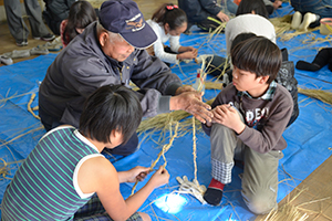 （写真）縄を編む児童とボランティアの方